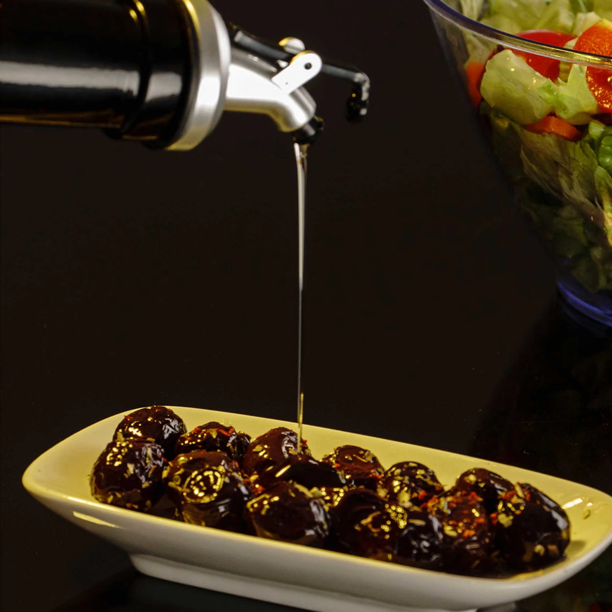 Oil being poured from the bottle over a dish of marinated olives beside a bowl of salad